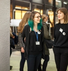 Zdjęcie. Dwie dzieczyny, wolontariuszki idą przez wystawę i rozmawiają. Dziewczyna po lewej ma zielone włosy do ramion i okulary. Dziewczyna po prawejma włosy do ramion. Obie ubrane na czarno, na szyi mają identyfikator Muzeum Sztuki Nowoczesnej.